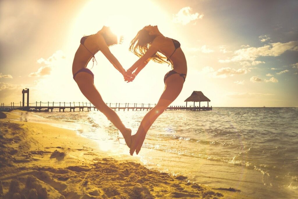 Zwei Frauen formen im Sprung ein Herz vor Sonnenuntergang am Strand - Sinnbild für weibliche Kraft, Verbindung und Lebensfreude
